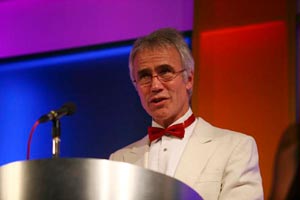 man in white dinner jacket speaking on stage