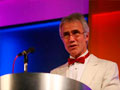 man in white dinner jacket speaking on stage
